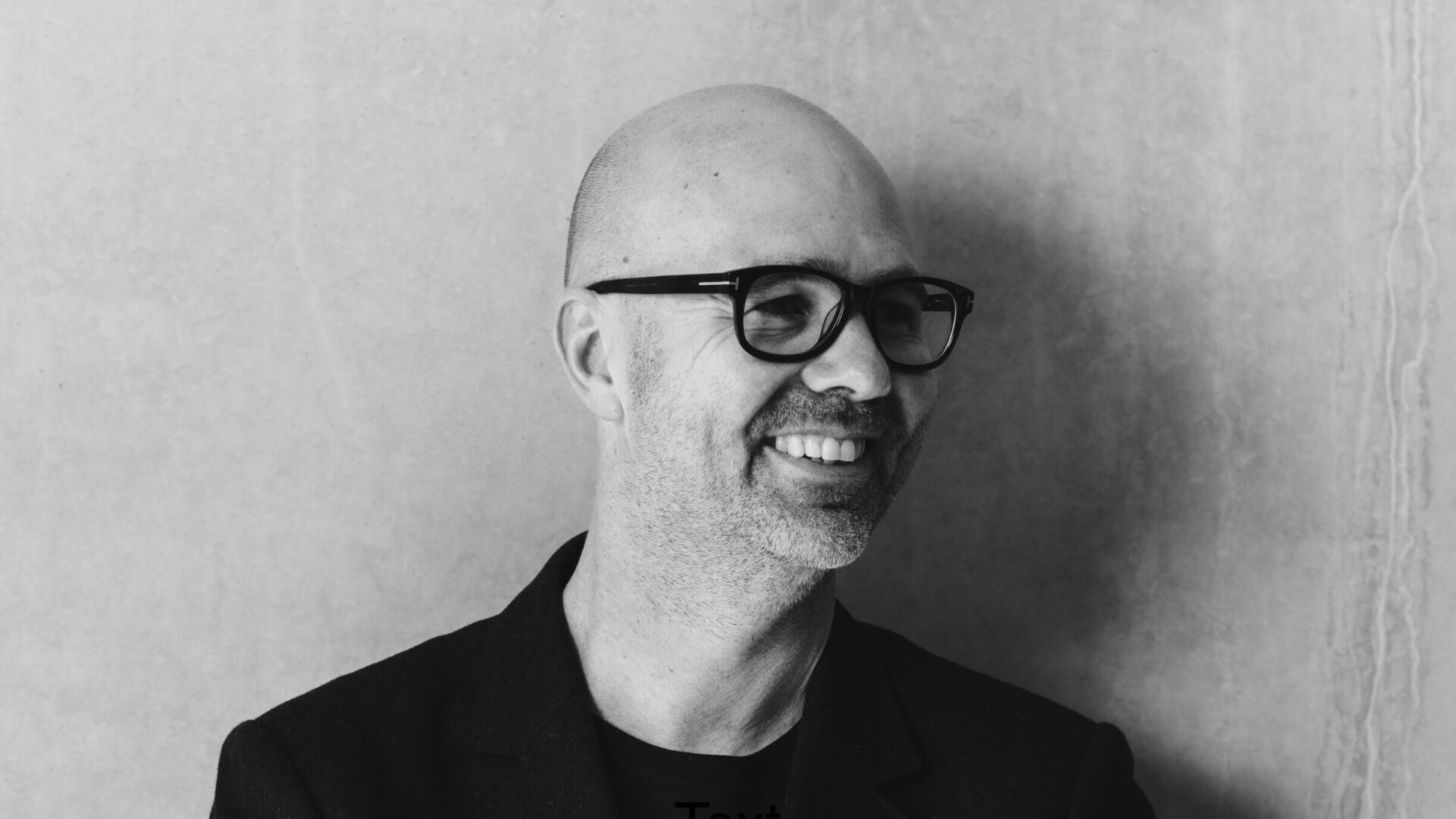 A portrait of architect Hamish Guthrie from Hecker Guthrie smiling and crossing his arms looking away from camera wearing a black jacket and t-shirt.